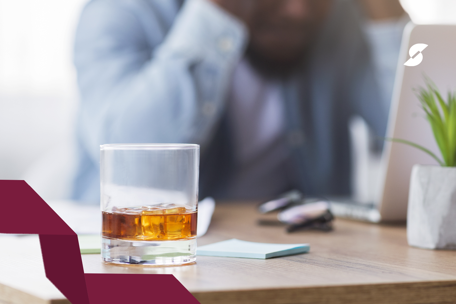 No foco da imagem temos um copo de bebida alcoólica em cima de uma mesa de trabalho. No fundo, desfocado, um homem com as mãos na cabeça, com expressão desanimada.
